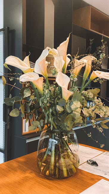 A large, elegant floral arrangement featuring white calla lilies with smooth, trumpet-shaped blooms and pale ivory petals, accompanied by delicate small white filler flowers and lush green foliage, in