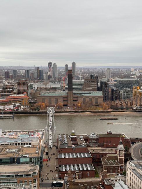 A panoramic view of London featuring a river in the foreground with boats sailing, modern skyscrapers, and historic buildings along the riverside, under a cloudy sky. The scene captures a mix of conte