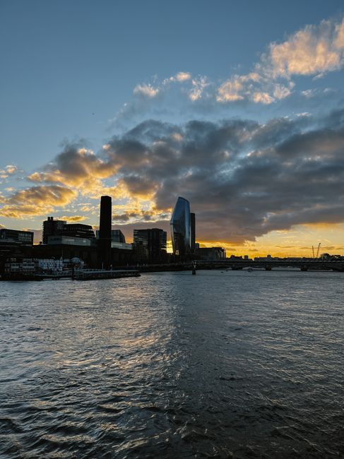 This image shows a view of the London skyline along the River Thames at sunset, featuring the modern, glass-clad skyscraper known as The Scalpel among other high-rise buildings. The sky is partly clou