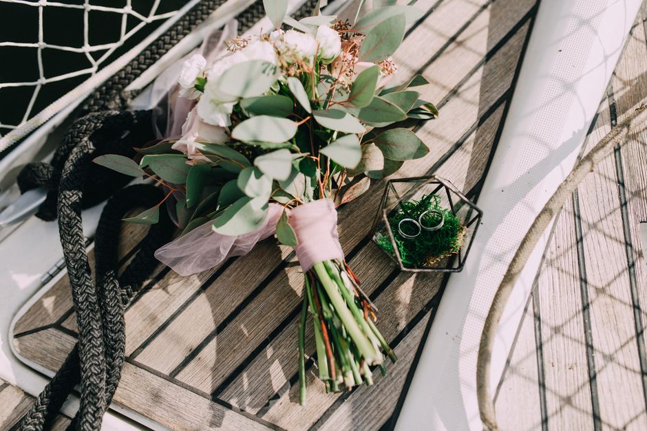 A bouquet of fresh white and blush pink flowers, including roses and eucalyptus, wrapped in soft pink paper with green stems visible, resting on a wooden surface. Nearby, a small glass terrarium with 