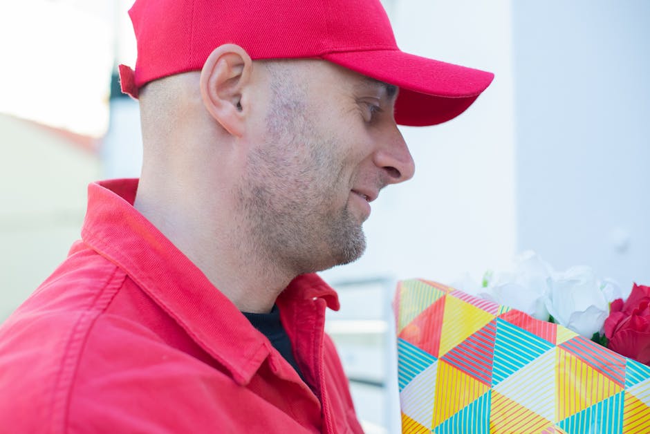 A delivery person wearing a red cap and matching red jacket is pictured holding a vibrant, geometrically patterned gift bag filled with fresh flowers, including roses in shades of pink and white. The 