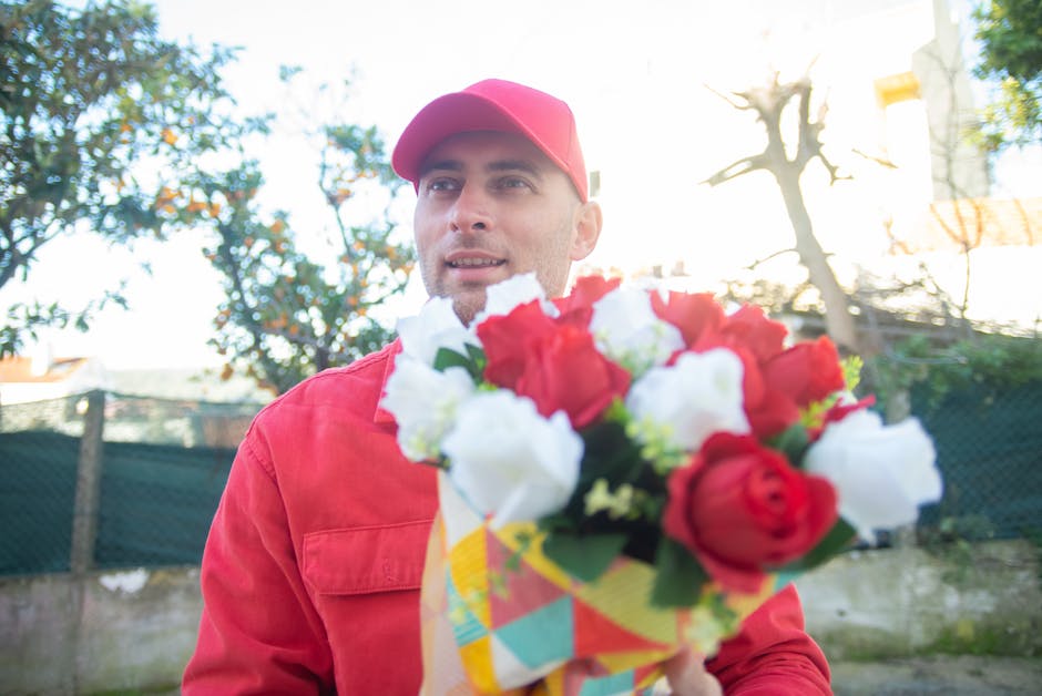 A man wearing a red cap and red jacket is holding a colorful, floral arrangement bouquet, featuring red and white roses, with green foliage. The bouquet is wrapped in decorative paper with geometric p